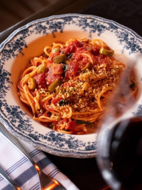 Plato de spaghetti con pomodoro, aceitunas verdes y pangrattato, servido en loza azul y blanca junto a una copa de vino tinto
